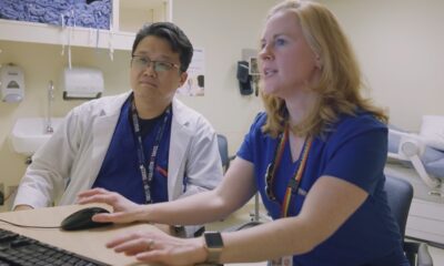 Two healthcare professionals, one wearing glasses and a lanyard and the other in blue scrubs, sitting at a desk and looking at a computer screen in a hospital setting.