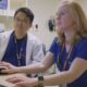 Two healthcare professionals, one wearing glasses and a lanyard and the other in blue scrubs, sitting at a desk and looking at a computer screen in a hospital setting.