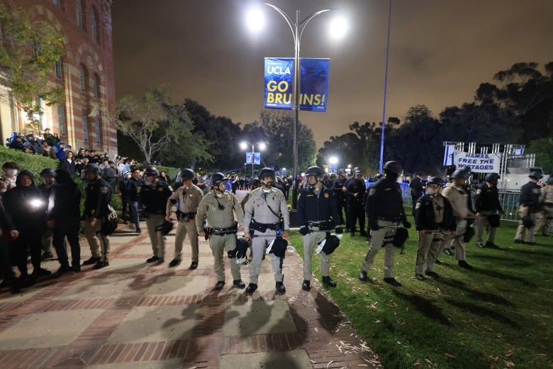 A line of police officers wearing helmets are shown standing on a university campus.