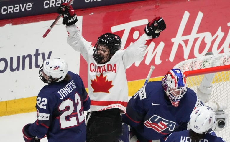 A female hockey player wearing red and white Canadian gear is seen celebrating a goal with both of her hands in the air.