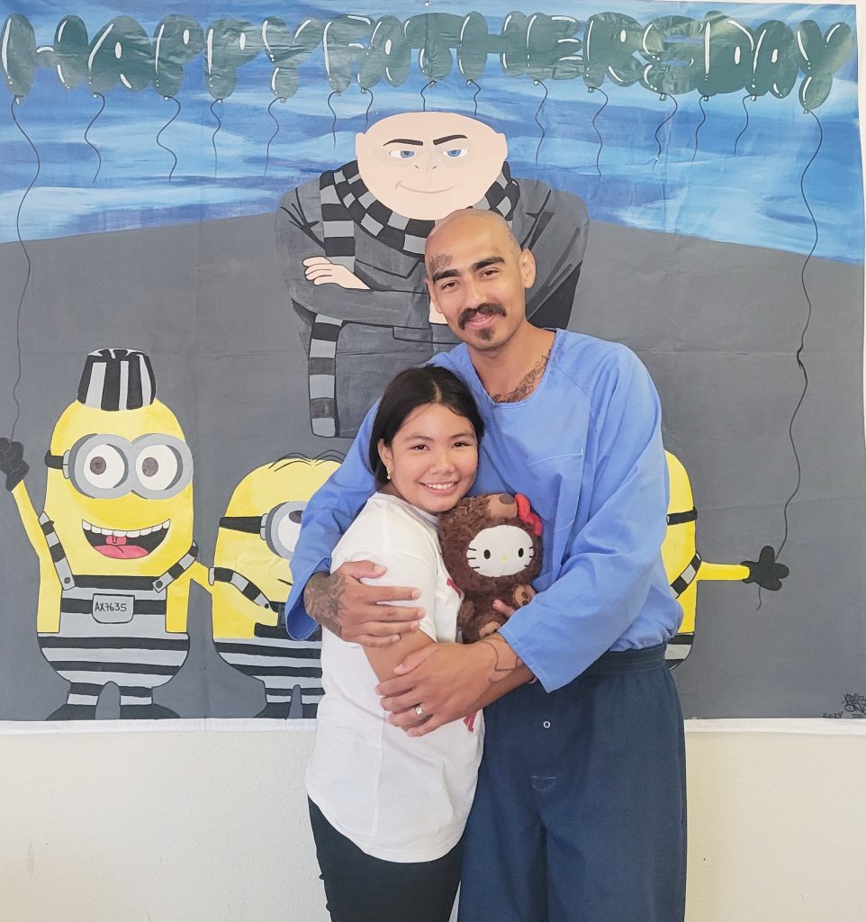 An incarcerated person hugs a visiting child in a prison visiting room.