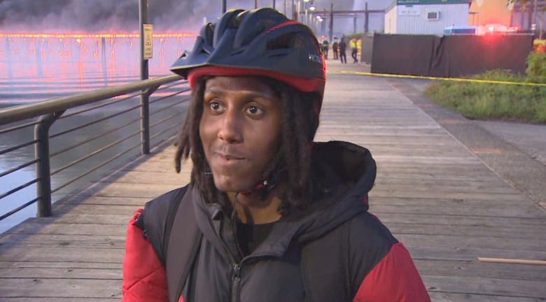 A man in a red bicycle helmet stands in front of a burning bridge. 