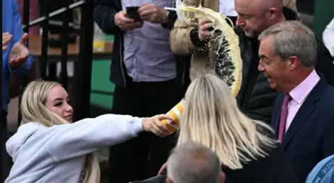 Getty Images Woman throwing milkshake in Nigel Farage's face. He is wearing a black blazer with a pink shirt an tie