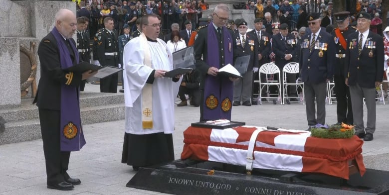 A man in white religious garment stands in front of a coffin draped in the Canadian flag.