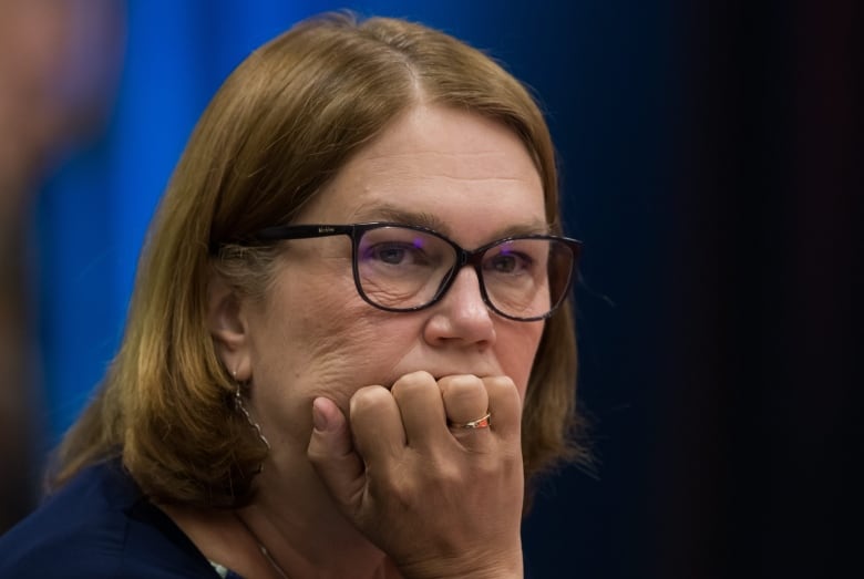 A woman with light brown hair and black glasses places her hand on her chin while looking seriously onwards.