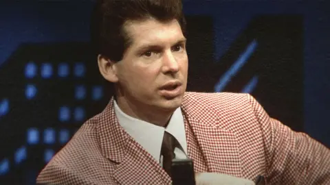 Mr. McMahon/Netflix A younger Vince McMahon from the 1980s or 1990s, wearing a checked pink and white suit with white shirt and black tie.