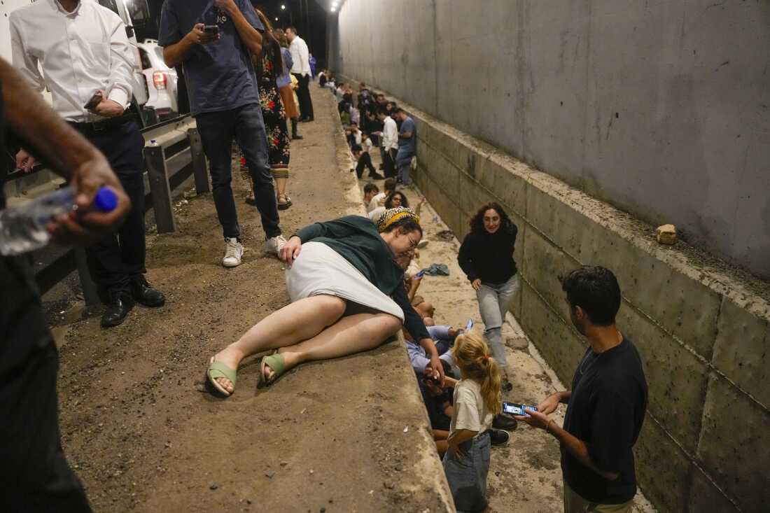 In this photo, people take cover by standing and sitting in a trench-like ditch on the side of a freeway in Shoresh, between Jerusalem and Tel Aviv. One of the freeway's guardrails appears on the left side of the photo.