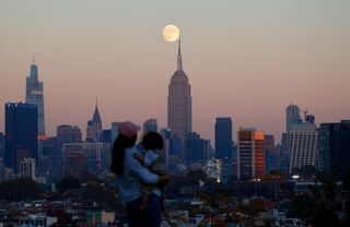 A large full moon in hovers just above the empire state building in a scene of the NYC skyline.