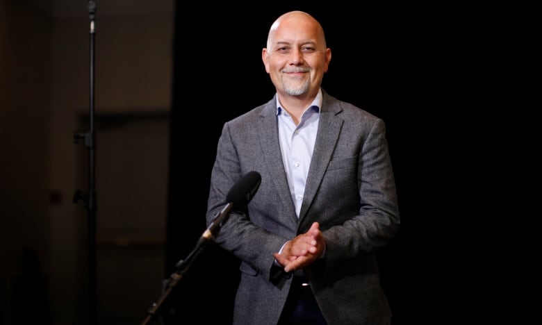 A bald Indigenous man with a white goatee wearing a grey jacket smiles while standing in front of a mic.