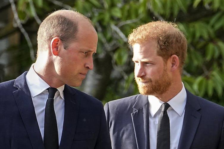 A tall bald man in a suit stands next to a tall bearded red-headed man.