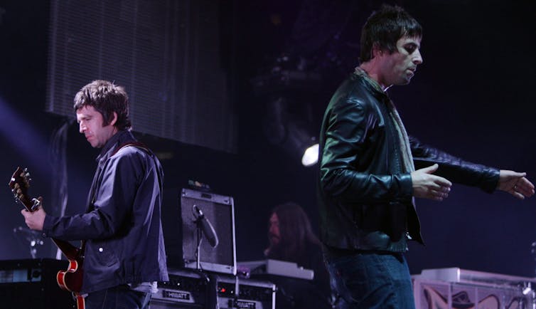 Two dark-haired men on a stage with their backs to one another. One holds a guitar.