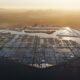 Aerial view of a futuristic, hexagonal water city with grid-like structures, surrounded by ocean and set against a desert landscape at sunset