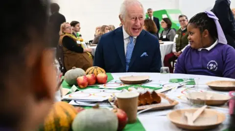 Getty Images A laughing Prince Charles seated at a table with a smiling schoolgirl wearing a purple school uniform sweatshirt to his left. On the table are plates with knives and forks and a collection of apples and various squashes. In the foreground is the blurred profile of another child.