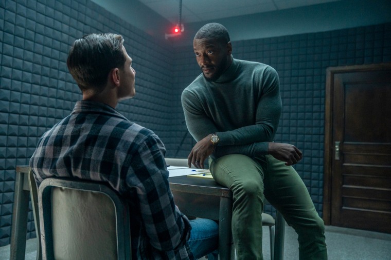 Aldis Hodge, right, sits on a table and questions someone sitting in front of him in a dark interrogation room