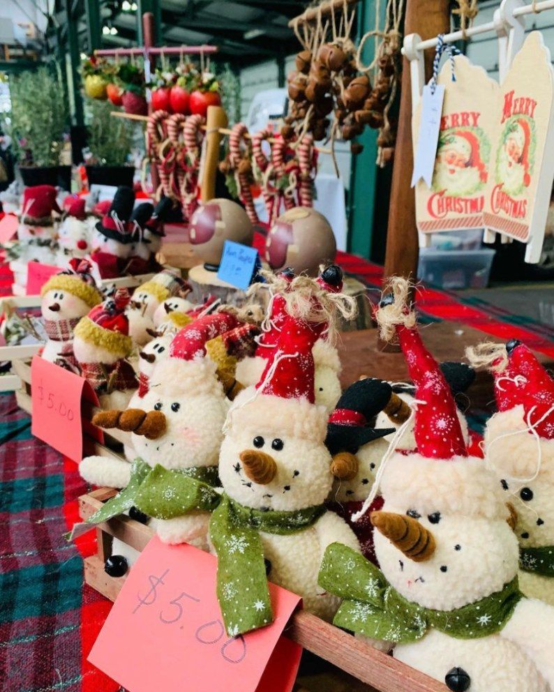 Stuffed snowman toys wearing festive hats are displayed for sale at a market stall, with prices labeled in front. Decorative items and garlic braids hang in the background.