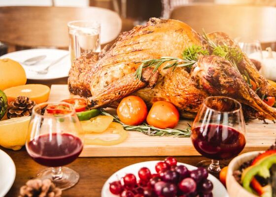 A roasted turkey garnished with herbs and surrounded by fruits and vegetables on a festive table, accompanied by two glasses of red wine.