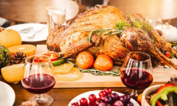 A roasted turkey garnished with herbs and surrounded by fruits and vegetables on a festive table, accompanied by two glasses of red wine.