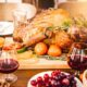 A roasted turkey garnished with herbs and surrounded by fruits and vegetables on a festive table, accompanied by two glasses of red wine.