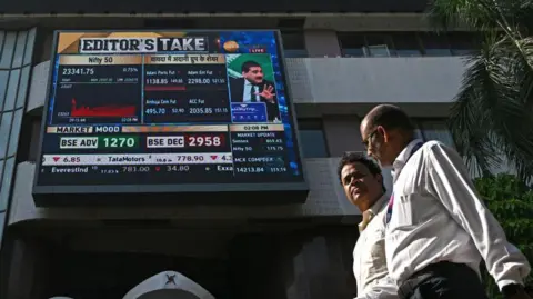 AFP Pedestrians walk past a digital broadcast on the facade of Bombay Stock Exchange (BSE) in Mumbai on November 21, 2024. Shares in Indian conglomerate Adani tanked on November 21 after its industrialist owner Gautam Adani was charged by US prosecutors with handing out more than $250 million in bribes for key contracts.