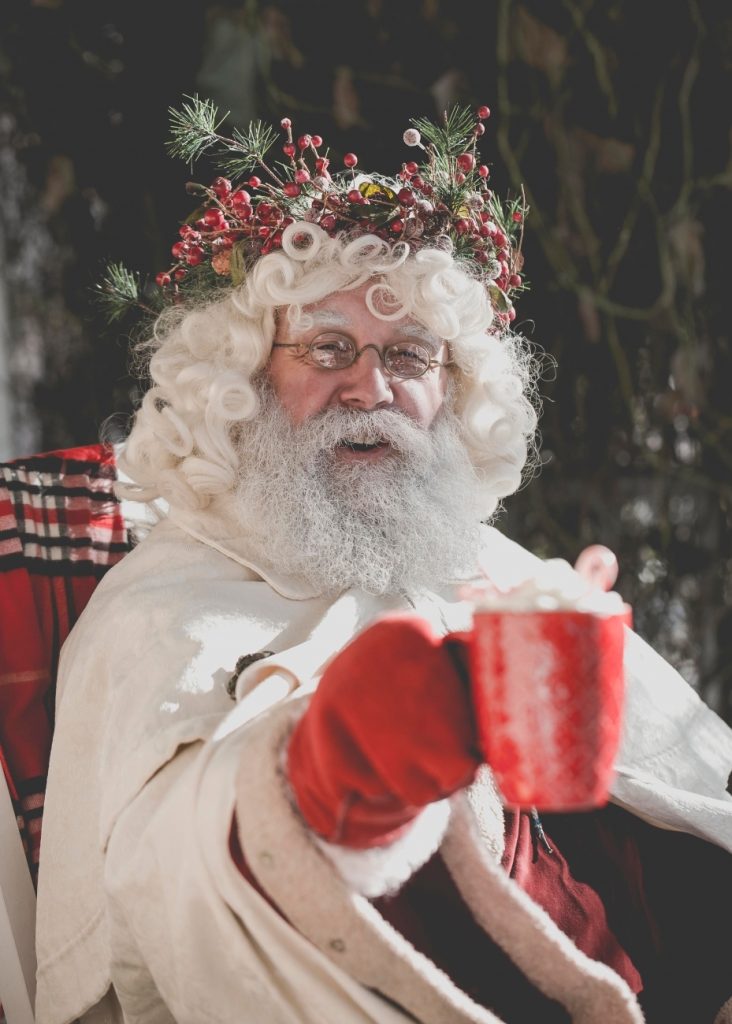 Santa Claus holding out a mug, wearing a festive wreath on the head, with a background of dark foliage.