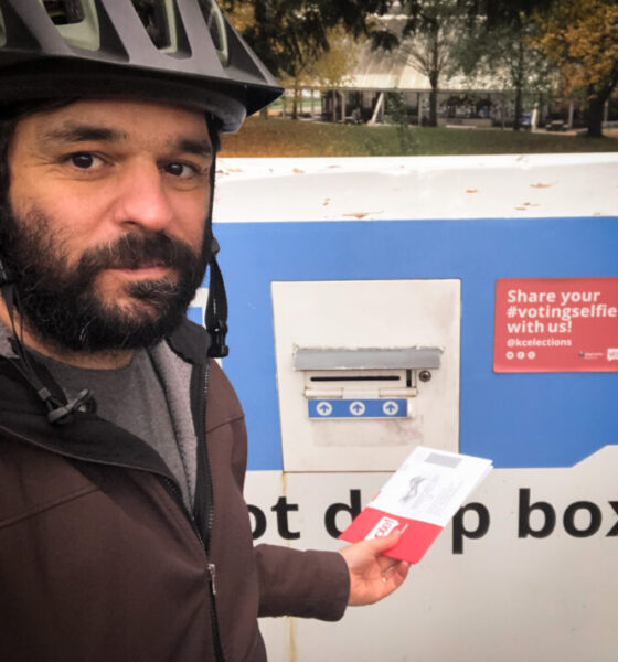 Selfie of the author wearing a bike helmet about to put a ballot into a drop box with gas works park in the background.