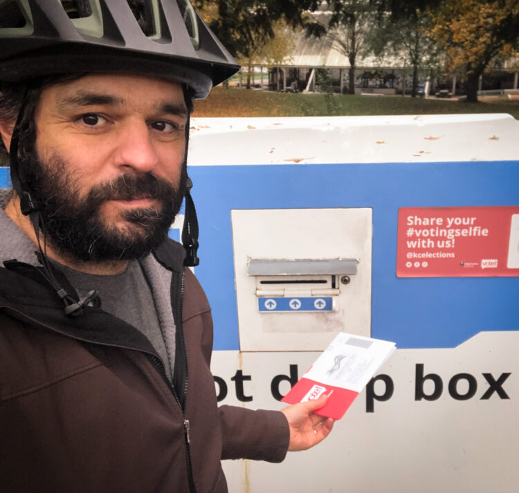 Selfie of the author wearing a bike helmet about to put a ballot into a drop box with gas works park in the background.