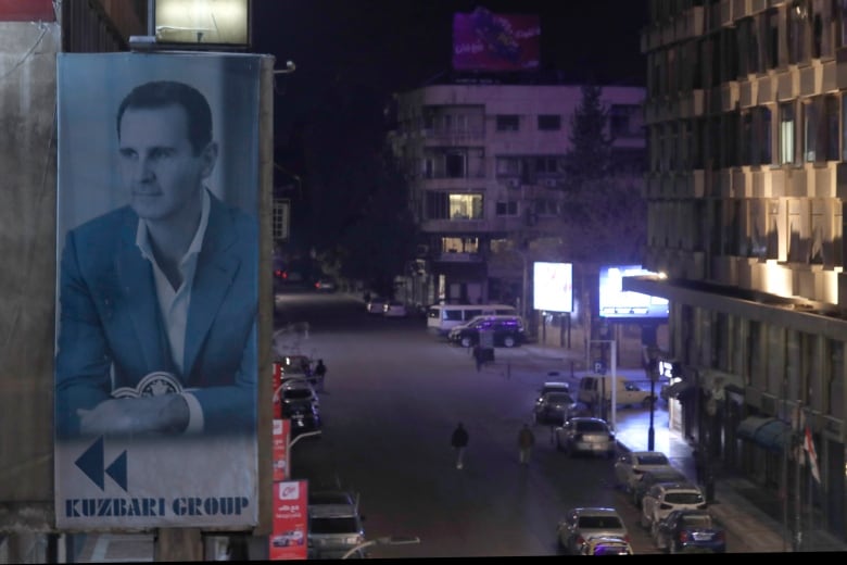 A giant banner featuring a portrait of a person hangs on the facade of a building as pedestrians and cars are seen on the street below.