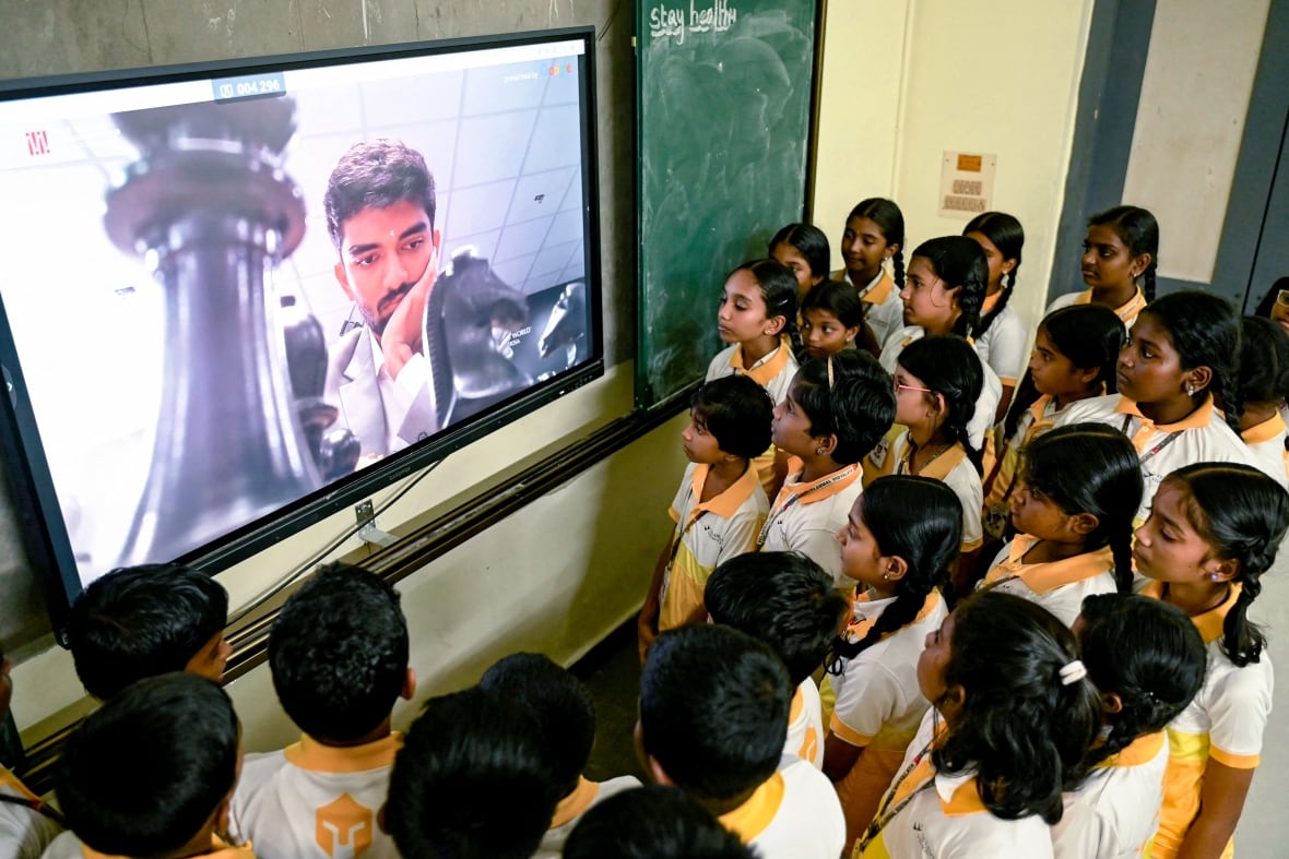 Several schoolchildren in uniform stand in front of a television set depicting a South Asian man and a giant chess piece.