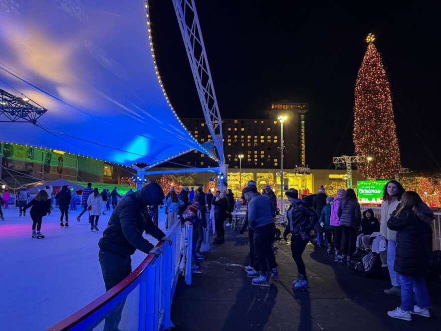 People stand off to the side of an ice rink where others skate. A tall Christmas tree stands in the background.