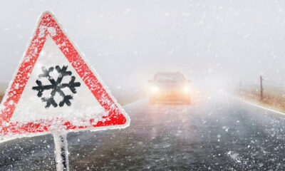 Snowfall warning sign surround by snow and a picture of a car with its headlights on on a highway.