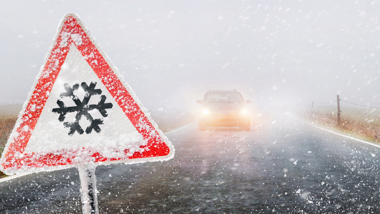 Snowfall warning sign surround by snow and a picture of a car with its headlights on on a highway.