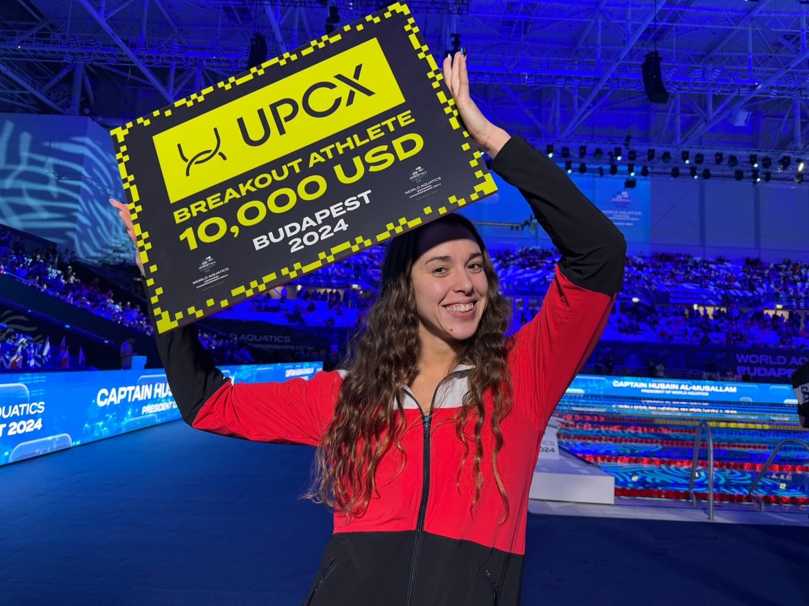 A woman smiles as she holds up a sign that reads 'Breakout Athlete, 10,000 USD, Budapest 2024.'