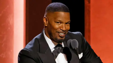 Getty Images Jamie Foxx is wearing a black tuxedo with a white shirt and is smiling at the camera