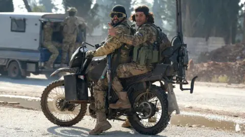 Getty Images Two men on a scrambler-style motorbike. Both are wearing desert-style camouflage uniforms and ammo belts. The driver has an AK-47 style rifle and there's a large machine gun strapped to the back of the bike. 