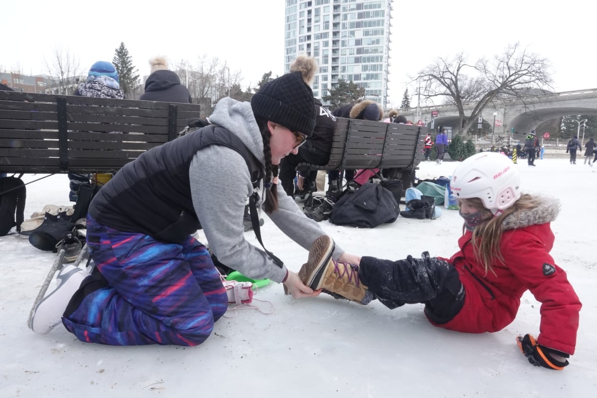 A child gets help with their boot on an outdoor skating rink.