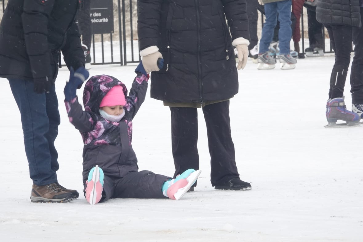 A child sits on the ice of an outdoor skating rink, holding the hands of two adults.