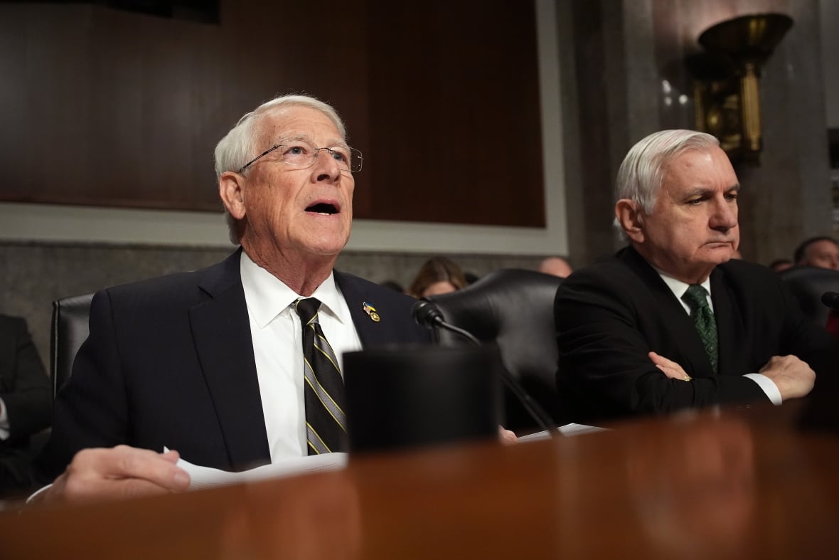 Two white haired, clean shaven men are shown wearing suits and ties seated at a long table. 