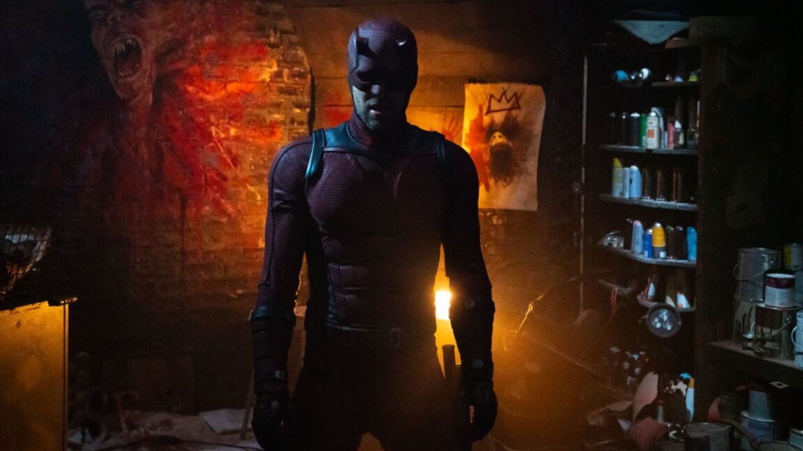 Charlie Cox in full Daredevil outfit in a dimly lit room with exposed brick wall