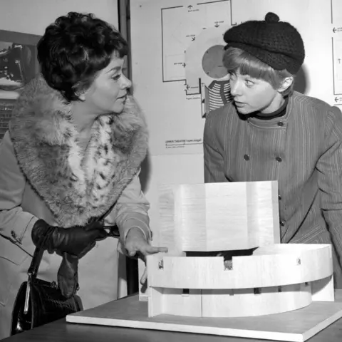 PA Media Joan Plowright and Geraldine McEwan talking and standing over a miniature model of the National Theatre, with architectural plans on the wall behind them